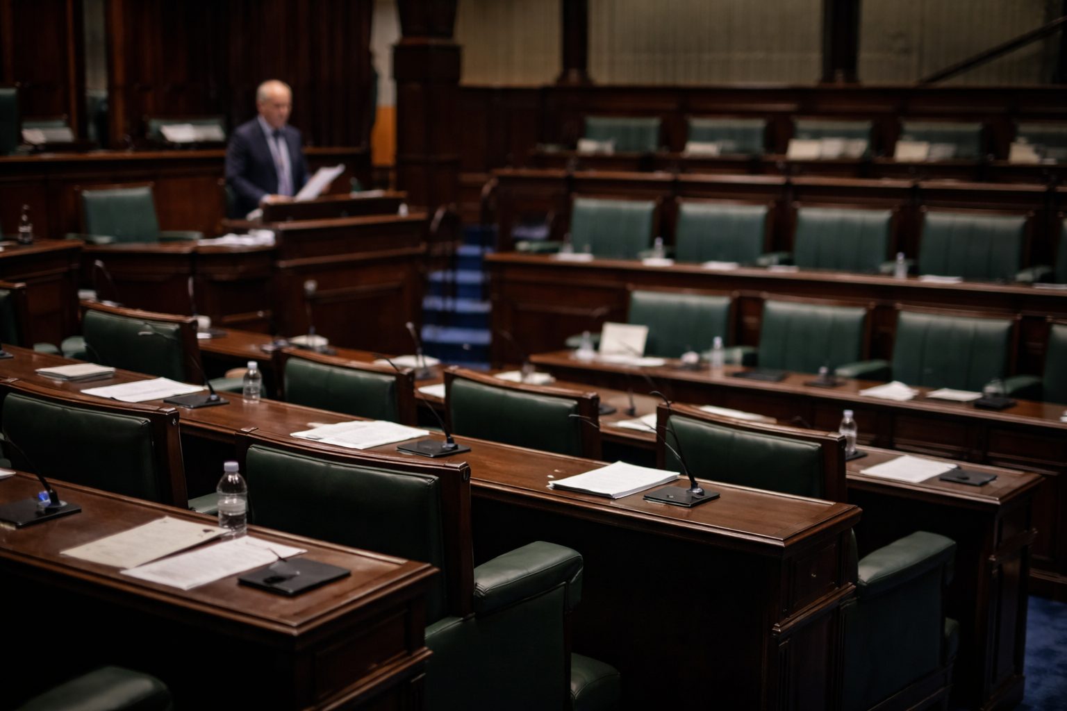 Irish Dáil chamber during a parliamentary debate, empty green seats and desks with a politician speaking in the background.